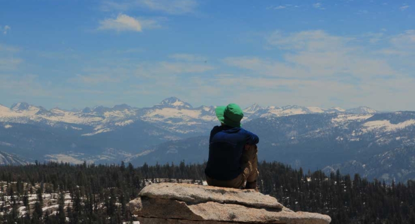A person sits on a rock with their back to the camera, overlooking the snow capped high sierra 